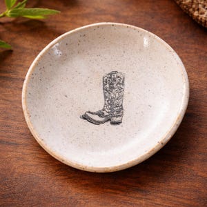 May include: A small, round, speckled ceramic dish with a hand-drawn image of a cowboy boot. The boot is black and white with intricate floral detailing. The dish has a slightly irregular shape and a rustic aesthetic.
