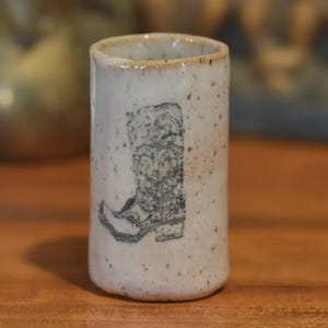 May include: A speckled, off-white ceramic tumbler with a black and gray illustration of a cowboy boot. The rim has a light brown glaze. The tumbler is cylindrical and appears handmade.
