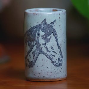 May include: A cylindrical ceramic tumbler with a speckled light blue-grey glaze. The exterior features a dark, etched illustration of a horse head in profile. The base of the vessel shows a thin band of exposed, unglazed reddish-brown clay.