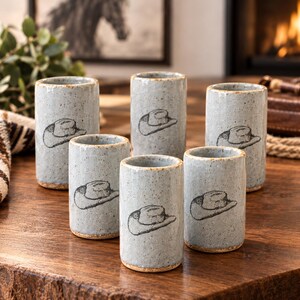 May include: A set of six cylindrical ceramic tumblers in a light blue speckled glaze. Each tumbler features a black line drawing of a cowboy hat on the front. The vessels have a smooth finish with a natural tan rim and base.