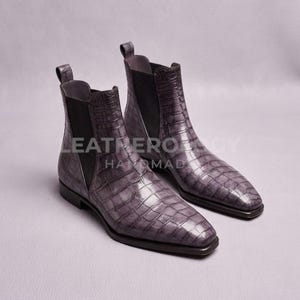 Peut inclure: Une paire de bottines Chelsea grises avec un motif texturé en peau de crocodile, des panneaux latéraux élastiques noirs et une languette au talon. Les bottes ont un bout pointu et un petit talon. Le texte LEATHEROLOGY HANDMADE est visible.