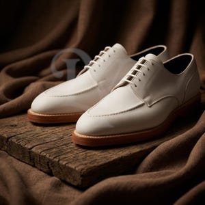 Peut inclure: Une paire de chaussures de ville en cuir blanc avec des semelles et des lacets marron clair. Les chaussures sont présentées sur une surface en bois vieilli, avec un fond en tissu marron.
