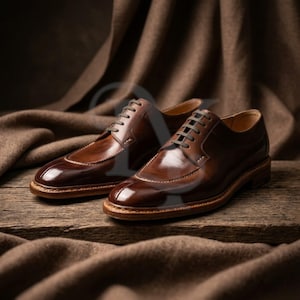 May include: A pair of polished brown leather dress shoes with a classic design. The shoes feature a cap toe, light-colored stitching, and brown laces. They are set on a wooden surface with a brown fabric backdrop.