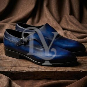 Könnte beinhalten: Ein Paar blaue Lederschuhe mit Schnallenverschluss. Die Schuhe werden auf einer Holzoberfläche vor einem braunen Hintergrund präsentiert. Das Leder hat einen Farbverlauf, der von Dunkelblau zu Hellblau übergeht.