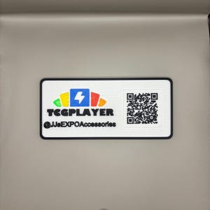 May include: A rectangular white sticker with a black border. The sticker features the text "TCGPLAYER" and "@JJsEXPOAccessories" in black. A colorful logo with a lightning bolt is on the left, and a QR code is on the right.