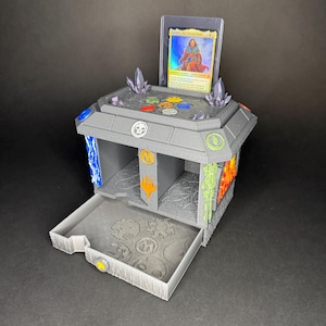 May include: A gray 3D-printed gaming card storage box featuring a pull-out drawer with embossed mana symbols. The top surface displays colorful mana icons and two crystal accents. A single trading card in a protective sleeve stands upright in the back.
