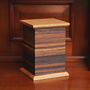 May include: A rectangular wooden urn with a light-colored top and base. The body of the urn is made of dark, striped wood. The urn is displayed on a wooden surface against a dark wooden background.
