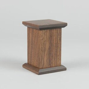 May include: A small, rectangular wooden pedestal with a dark brown finish. The pedestal has a square top and base, with a textured, vertical grain pattern on the sides. The design is simple and elegant, suitable for displaying small objects.