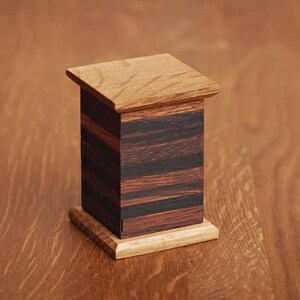May include: A small, wooden box with a layered design. The box features a light-colored base and top, with a dark, striped body. The wood grain is visible, and the box has a simple, geometric shape. The box is likely a decorative item or a small storage container.