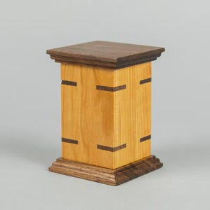 May include: A handcrafted wooden box with a rectangular shape. The box is made of light-colored wood with dark brown accents and a dark brown top. The box has a decorative base and is likely used for storage.