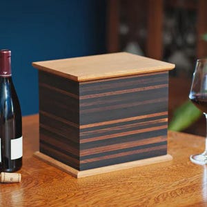 May include: A wooden box with a light-colored top and base, and a dark, striped body. A wine bottle, a corkscrew, and a wine glass with red wine are on a wooden table. The background is a dark blue wall.