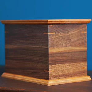 May include: A handcrafted wooden box with a rectangular shape. The box is constructed from dark brown and light brown wood, with visible grain patterns. The corners are joined with contrasting wood accents. The box sits on a wooden surface against a blue background.