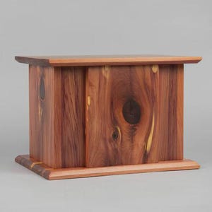 May include: A rectangular wooden box with a natural wood grain finish. The box is made of reddish-brown wood with a lighter-colored top and base. The wood grain is prominent, with knots and variations in color. The box is likely a decorative storage container.