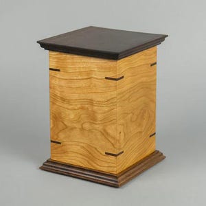 May include: A wooden pedestal with a dark brown top and base. The main body is light-colored wood with a visible grain, accented by dark brown inlays. The pedestal is rectangular and stands on a gray background.