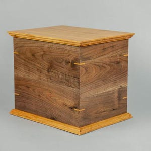 May include: A rectangular wooden box with a light brown top and base, and a darker brown body. The box is constructed with visible wood grain and small, light-colored accents at the corners.