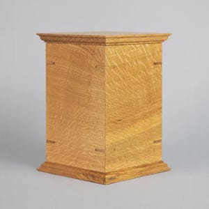 May include: A light brown wooden pedestal with a square top and base. The pedestal is constructed with visible joinery and has a natural wood grain pattern. The design is simple and elegant, suitable for display.
