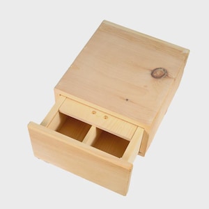 May include: A light-colored wooden box with a partially open drawer. The drawer is divided into two compartments. The wood grain is visible, and the box has a natural, unfinished appearance. The box is likely for storage.