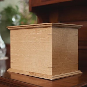 May include: A light-colored wooden box with a rectangular shape. The box has a detailed, layered top and base. The wood grain is visible, and the box is set on a dark wooden surface. The background includes a glass decanter and a wooden cabinet.
