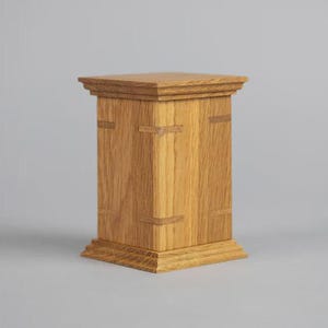 May include: A rectangular wooden box with a light brown finish. The box has a square top and base, with decorative trim. The sides of the box are adorned with rectangular accents. The box is made of wood and has a natural grain.