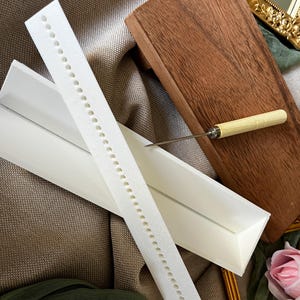 May include: A white foam strip with evenly spaced holes, a white triangular box, a wooden block, and a small awl with a light-colored wooden handle. The items are arranged on a textured, neutral-toned surface.