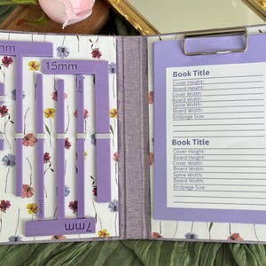 May include: A purple bookbinding tool set featuring various guides with labelled measurements of 6mm, 7mm, and 15mm. The set is organised on a floral patterned board inside a folder, which includes a clipboard with a printed book title form.