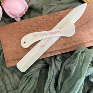 May include: Two white bone folders with a smooth, curved design and a pointed tip. Each tool features the text The Cozy Bindery printed in pink lettering. One folder includes a small circular hole at the rounded end for hanging or storage purposes.