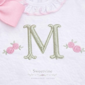 Pode incluir: Body de bebé branco com uma letra "M" bordada em verde claro e dois grupos de rosas cor-de-rosa. Um grande laço rosa está no canto superior esquerdo. O body tem uma gola com babados.