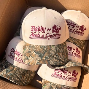 May include: A cream-colored baseball cap with a camouflage brim. The front features maroon embroidered text that reads "Daddy Needs a Cigarette" alongside a small graphic of a cigarette pack. The cap has a structured crown and a button on top.