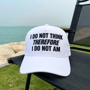 May include: A white baseball cap featuring the black text I DO NOT THINK THEREFORE I DO NOT AM printed in a bold, sans-serif font across the front panel. The cap has a structured crown, a curved brim, and a button at the top center.