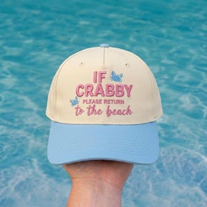 May include: A cream-colored baseball cap with a light blue brim. The front features embroidered pink text that reads IF CRABBY PLEASE RETURN to the beach, accompanied by two small light blue crab icons positioned near the text.