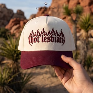 May include: A cream-colored baseball cap with a dark maroon brim and matching maroon embroidery on the front. The embroidery features the text "hot lesbian" in a stylized gothic font, framed by flame-like shapes. Above the cap, the text "K THE EXCLUSIVE!" appears with a small star icon.
