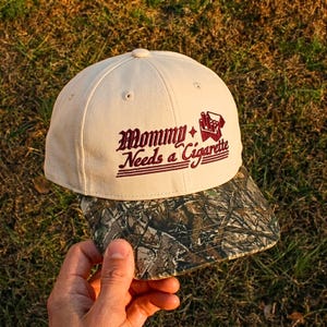May include: A cream-colored baseball cap with a camouflage-patterned brim. The front features dark red embroidered text that reads Mommy Needs a Cigarette, accompanied by a small graphic of a cigarette pack with a four-pointed star icon.