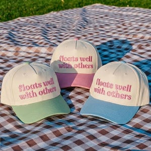 May include: Three baseball caps with off-white crowns and colored brims in light green, pink, and light blue. Each cap features the embroidered text "floats well with others" in a pink, rounded, serif font across the front panel of the crown.