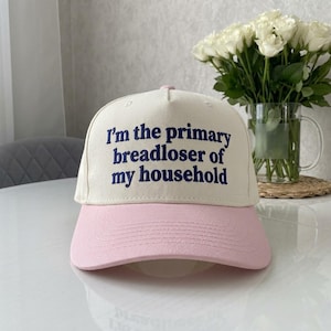 May include: A cream-colored baseball cap with a pink brim. The front panel features dark blue embroidered text that reads, I'm the primary breadloser of my household. The cap has a structured crown with a button on top and visible stitching.