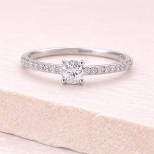 May include: A silver ring with a large, round, clear gemstone in the center. The band is lined with small, clear gemstones. The ring is set against a light-colored background.