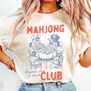May include: Ivory-colored t-shirt with a red "MAHJONG CLUB" graphic. The graphic features a table with a Mahjong game and four dogs playing. The text "dragons, dogs & drink" is below the table.
