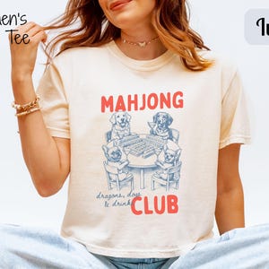 May include: Ivory boxy tee with a graphic of dogs playing mahjong. The text "MAHJONG CLUB" is in red, with "dragons, dogs & drinks" below. The shirt has short sleeves and a crew neck.