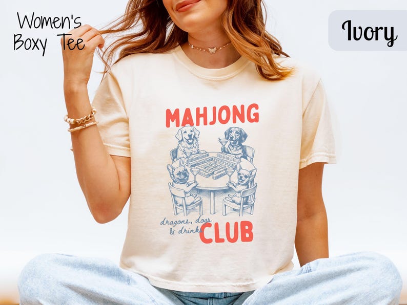 May include: Ivory boxy tee with a graphic of dogs playing mahjong. The text "MAHJONG CLUB" is in red, with "dragons, dogs & drinks" below. The shirt has short sleeves and a crew neck.