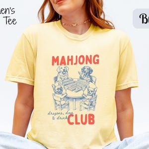 May include: Pale yellow boxy tee featuring a dog-themed mahjong graphic. The text reads "MAHJONG CLUB" with "dragons, dogs & drink" below. The shirt is a light yellow colour.