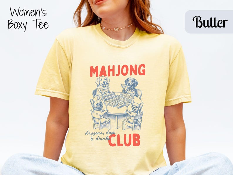 May include: Pale yellow boxy tee featuring a dog-themed mahjong graphic. The text reads "MAHJONG CLUB" with "dragons, dogs & drink" below. The shirt is a light yellow colour.
