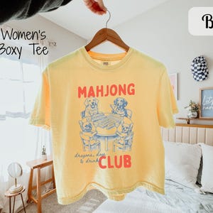 May include: A yellow boxy tee shirt featuring a graphic of dogs playing mahjong. The text "MAHJONG" is above the dogs, and "Dragons, dogs & drink CLUB" is below. The shirt is suspended from a wooden hanger.
