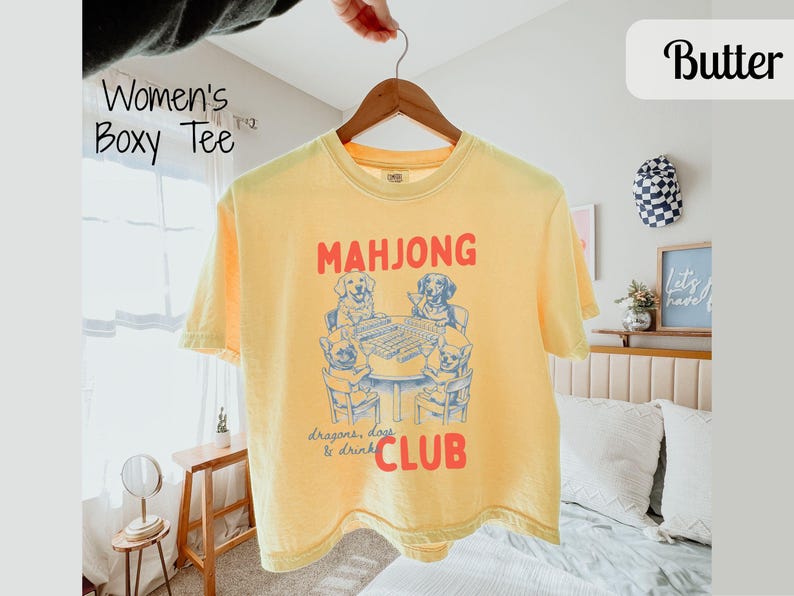 May include: A yellow boxy tee shirt featuring a graphic of dogs playing mahjong. The text "MAHJONG" is above the dogs, and "Dragons, dogs & drink CLUB" is below. The shirt is suspended from a wooden hanger.