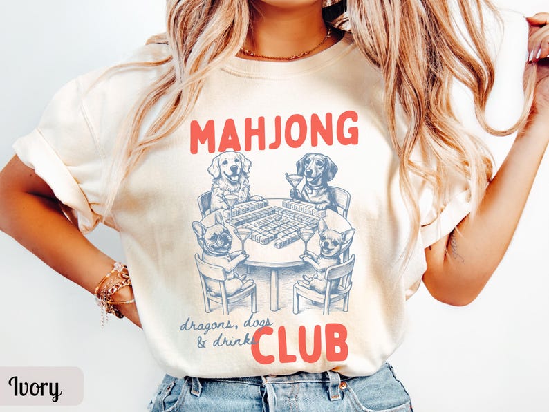 May include: Ivory-colored t-shirt with a red "MAHJONG CLUB" graphic. The graphic features a table with a Mahjong game and four dogs playing. The text "dragons, dogs & drink" is below the table.