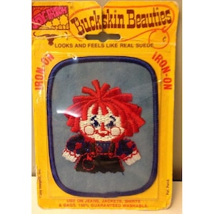 May include: A rectangular iron-on patch featuring an embroidered doll with bright red hair, a blue and white outfit, and rosy cheeks, centered on a light blue background. The packaging is yellow with the text Buckskin Beauties and Joy Patch.