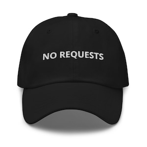 May include: Black baseball cap with the embroidered text "NO REQUESTS" in white. The cap has a curved brim and a button on top. A casual accessory for everyday wear.