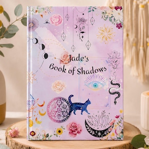 Puede incluir: Un libro de tapa dura con una cubierta de color morado claro que presenta el texto "Jade's Book of Shadows". El diseño incluye ilustraciones de un gato azul, rosas, un sol, una luna, una serpiente, un ojo, cristales y motivos florales.
