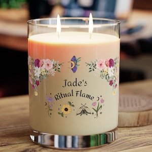 Puede incluir: Una vela grande de dos mechas en un tarro de cristal transparente. La vela tiene un degradado de colores melocotón y crema. Ilustraciones florales y celestiales rodean el texto "Jade's Ritual Flame". La vela está encendida, con dos llamas visibles.