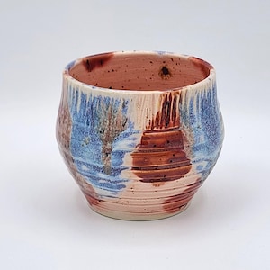 Può includere: Una tazza in ceramica fatta a mano con un design unico e strutturato. La tazza presenta una miscela di colori, tra cui sfumature di blu, marrone e crema. L'interno è di un caldo marrone rossiccio. L'esterno ha un effetto a strati, con motivi gocciolati e a spirale.