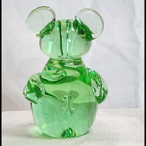 May include: A green glass bear figurine with large, rounded ears and small arms. The bear is made of transparent green glass, with visible bubbles and swirls within the glass. The figurine has a smooth, rounded body and is displayed on a white surface.