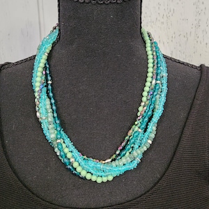 May include: A multi-strand necklace with beads in shades of turquoise, teal, and green. The necklace is displayed on a black mannequin. The beads vary in size and texture, creating a layered effect. Some beads have a metallic sheen.